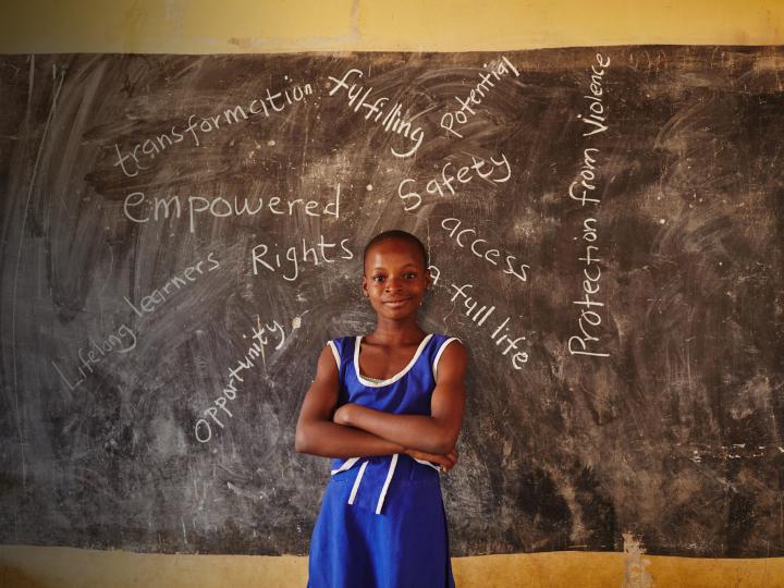 A young girl stands in her school with words written on a blackboard