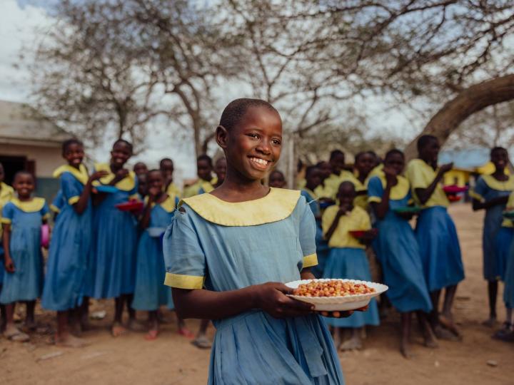 A smiling girl in a blue and yellow school uniform holds a plate of food outdoors, with classmates in matching uniforms gathered behind her