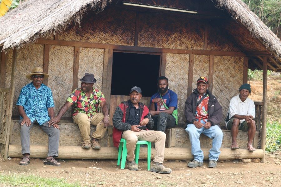 A group of respected community leaders from Sepionda, Lower Mendi District, Southern Highlands Province, sit in a straight row in front of a Hausman (a men's house). Pictured are David Onge, Ward Councillor Richard Pipi, Community Leaders Nelson Embisi and David Pone, and a young man named Dream. The respect for the elders is visible through the nods and murmurs of the younger men gathered around them. Ward Councillor Richard Pipi, wearing a PNG cap, is the first to speak, addressing the small crowd assembled at Community Leader David Pone's residence.