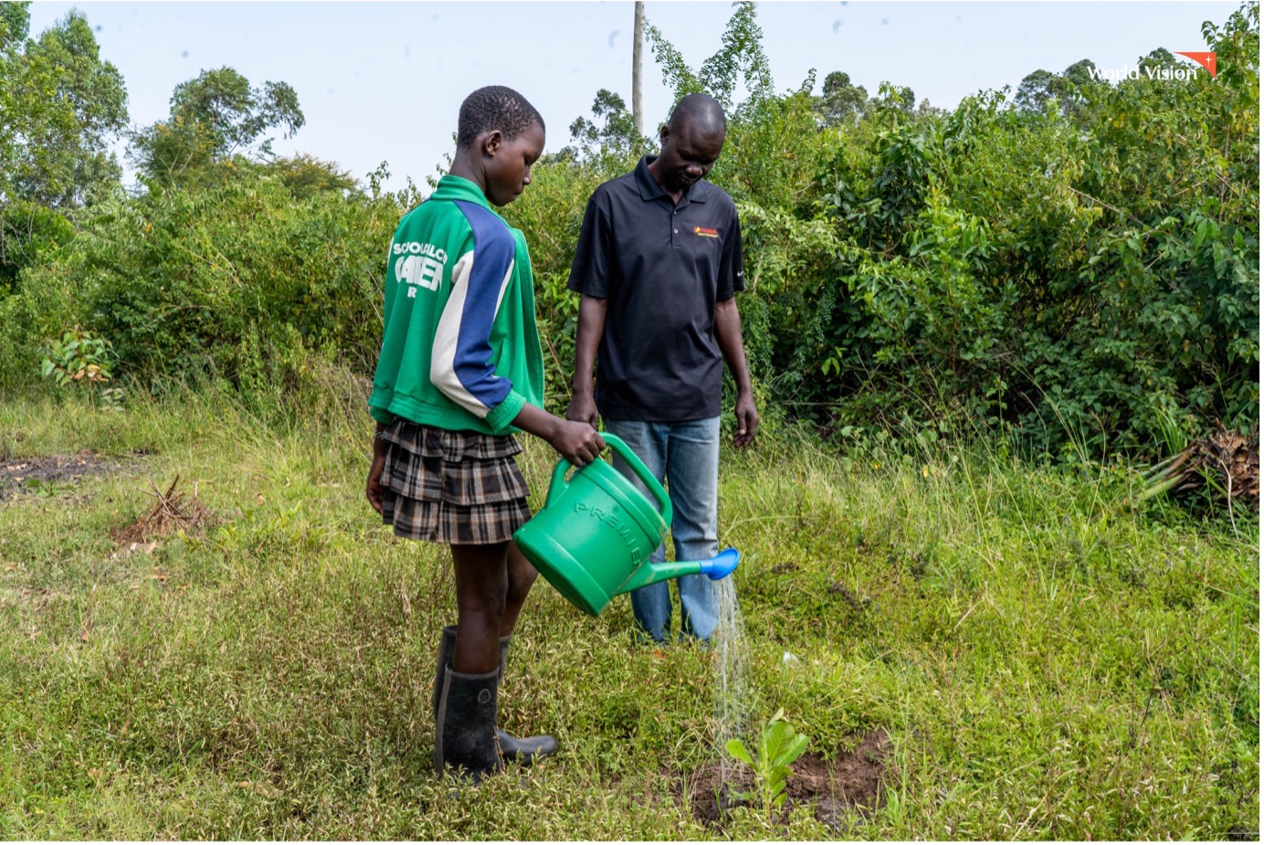 A young girl named Elizabeth waters trees with a watering can, guided by her father, who stands nearby offering support.