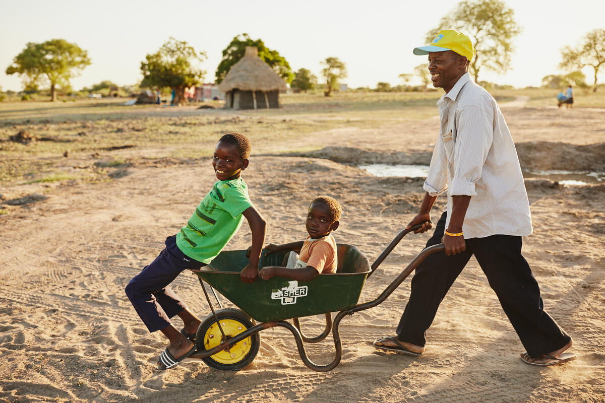 A man playing and pushing a wheelbarrow with two children in it