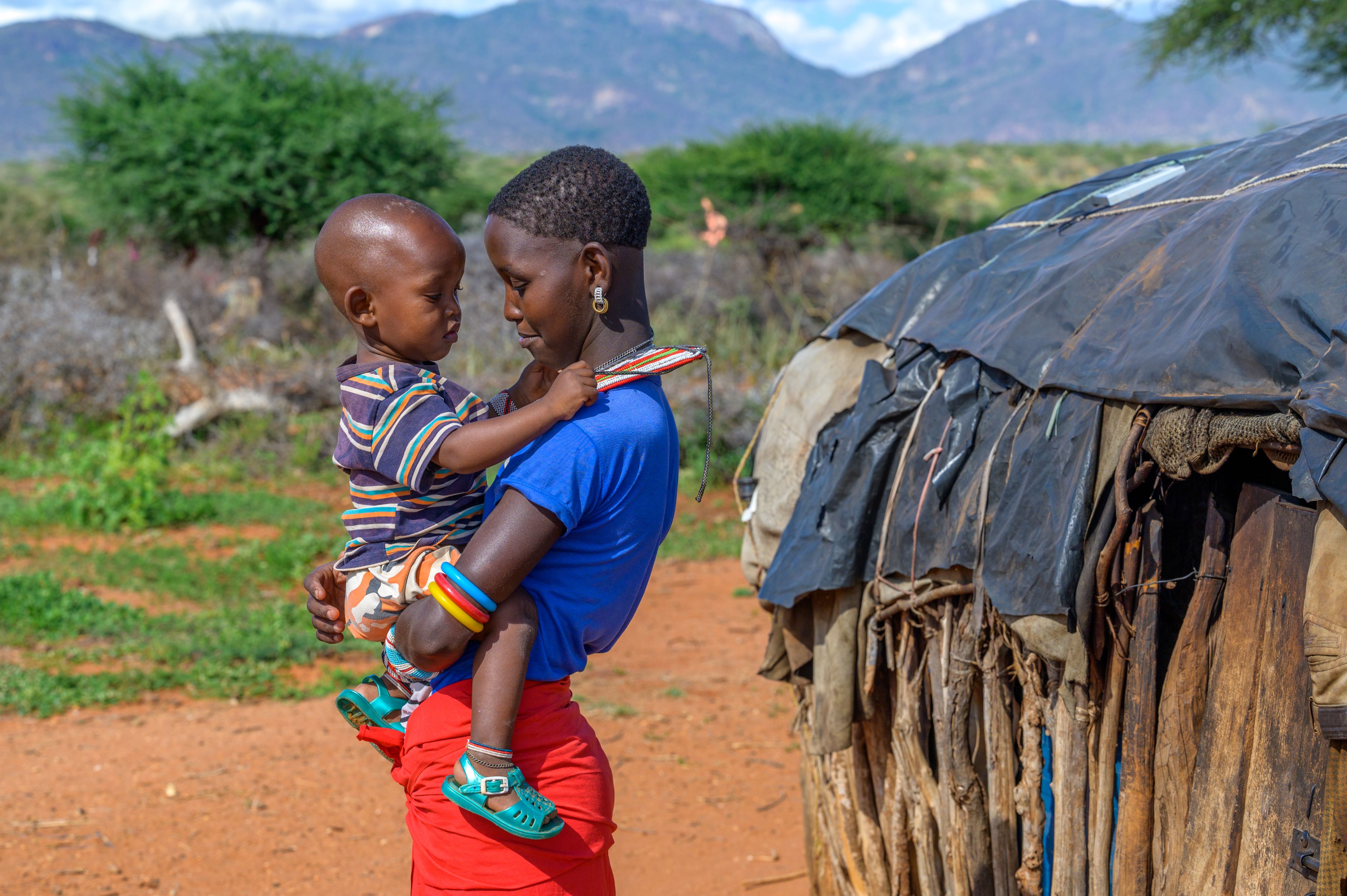 A young woman in a bright blue shirt holds a young child close, standing outside near a traditional hut, with greenery in the background.