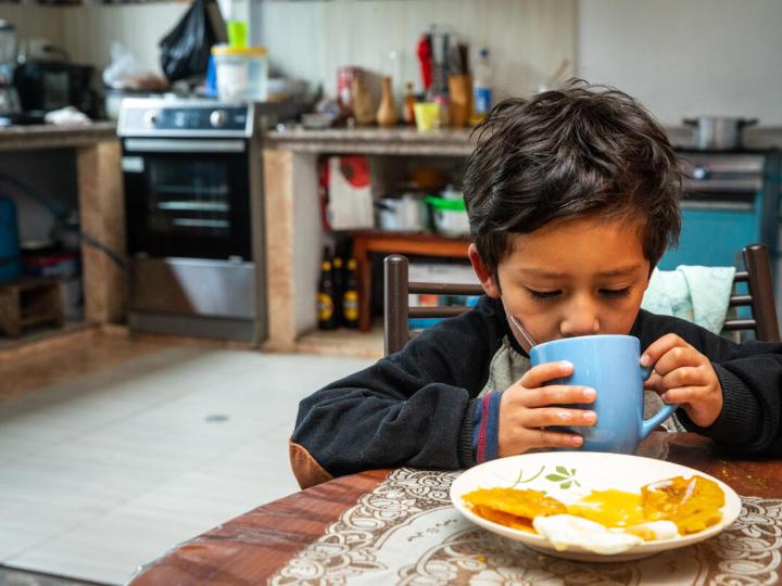 young boy drinks out of a cup at a kitchen table and a plate of food sits in front of him
