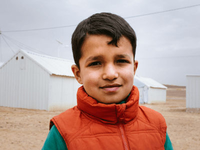 Syrian refugee child wearing an orange gilet in front of a white tent