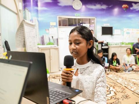 A young girl in Indonesia holds a computer and microphone to present to a group of adults