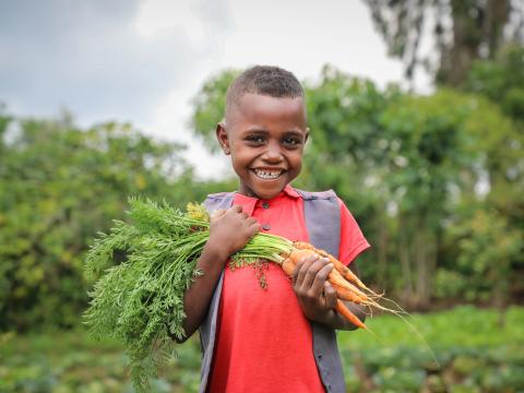 Kifle's child proudly holds a freshly picked cabbage and carrot from their garden.