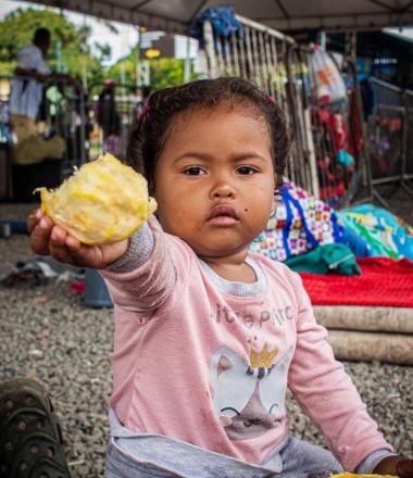 child sitting on the ground holding up a piece of food. 